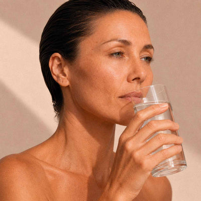Femme buvant un verre d'eau sur fond beige, geste d'hydratation quotidienne