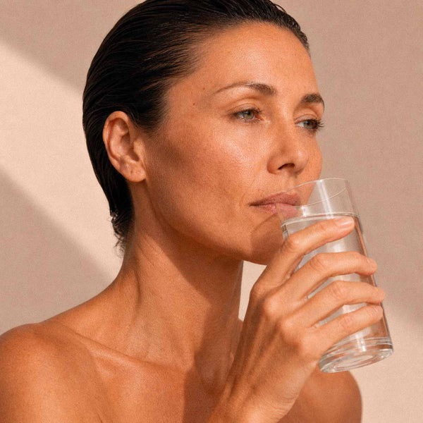 Femme buvant un verre d'eau sur fond beige, geste d'hydratation quotidienne