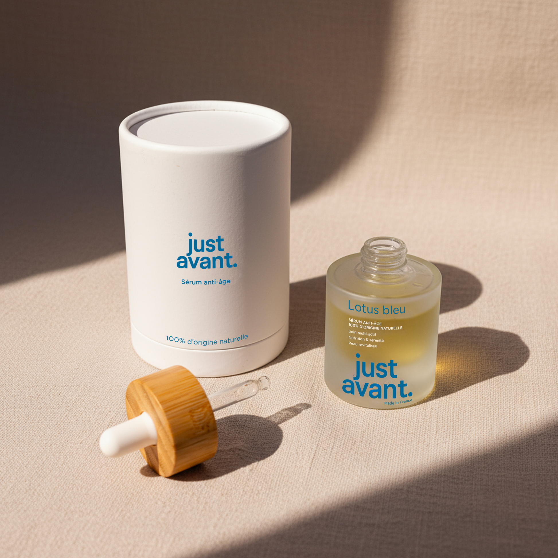 Packshot du sérum visage Lotus Bleu Just Avant avec flacon en verre givré, pipette en verre à bouchon en bois et étui cylindrique blanc, présenté sur fond textile clair.